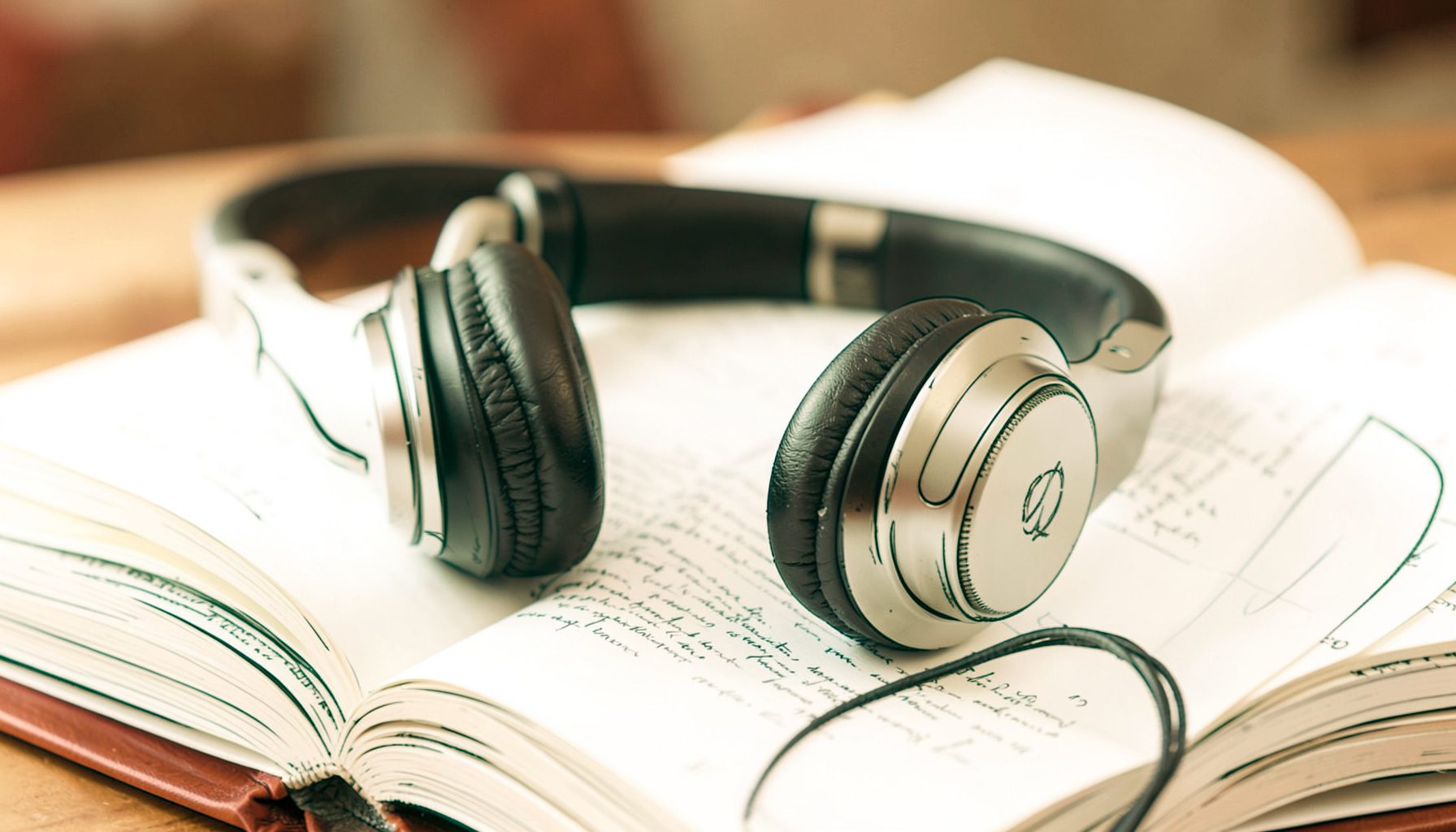A warm, softly lit close-up of a pair of over-ear headphones resting on an open hardcover book, with deep browns, muted golds, and cream tones creating a quiet literary atmosphere.